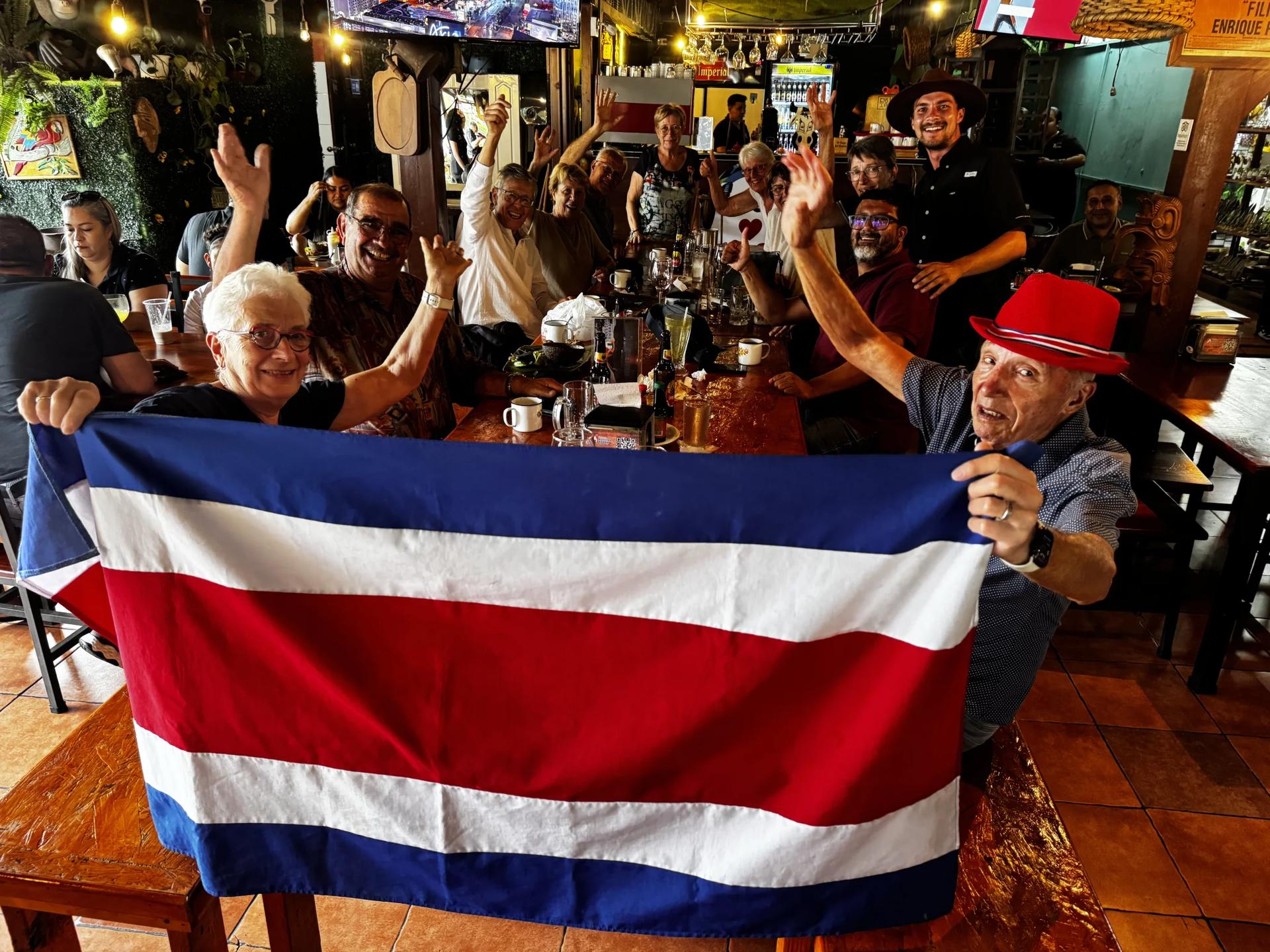 Groupe de voyageurs lors d'un circuit au Costa Rica avec Vibralma Travel autour d'une table dans un restaurant, souriants et levant le drapeaux du Costa Rica et accompagnés du guide expert naturaliste Lucas Boyat